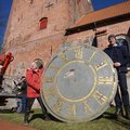 Eine ältere Frau und ein Mann mittleren Alters präsentieren ein großes Ziffernblatt einer Kirchturmuhr.